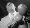 A dark-skinned man wearing a light-colored suit and tie and holding two maracas is seen from below at an angle