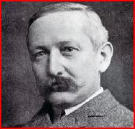 A head and shoulders view of a man with thinning hair and a moustache. He is wearing a suit jacket and tie.