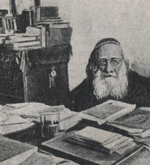 Photographie noir et blanc d'un homme agé portant des lunettes et une barbe blanche. Il est assis devant sa table de travail où se trouvent de nombreux livres.