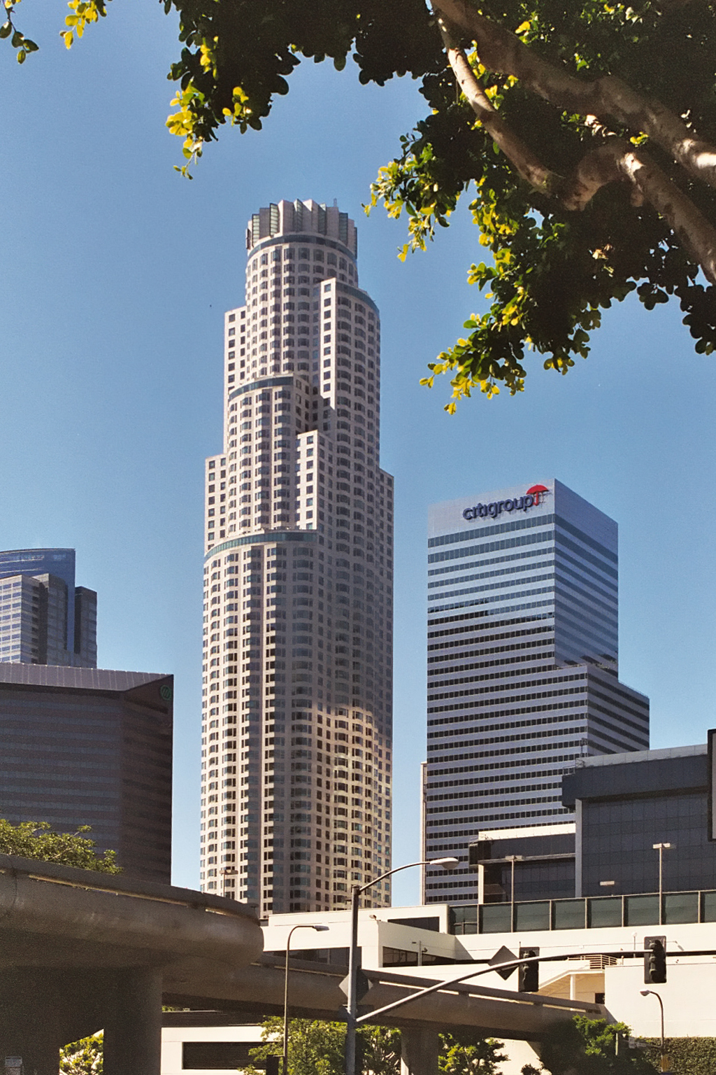 Framed by a tree branch, several tall buildings can be seen, the tallest, and centered, building looks somewhat like a wedding cake, made of three oval cylinders one on top of the other