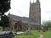 Gray stone building with ornate square tower and slate roof. In the foreground are gravestones.