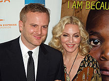 A blond middle-aged woman and a middle-aged man stand beside each other, smiling towards the camera. The woman wears a brown caftan while the man is in black suit.