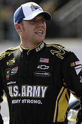 A man in his twenties wearing a white and blue baseball cap, and a black racing suit.