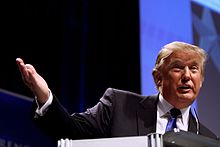 Donald Trump, dressed in a black suit with white shirt, and blue tie. He is facing toward the viewer and speaking at the Conservative Political Action Conference in February 2011.