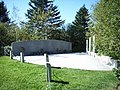 Photo du mémorial, comportant un sol en pierre en forme de triangle avec un mur en pierre assez bas de plusieurs mètres de long situé sur un des côtés du sol. Le petit espace est entouré d'herbes, au premier plan, et d'arbres à l'arrière-plan. Des petits poteaux reliés par une chaîne délimitent le contour de l'espace commémoratif.