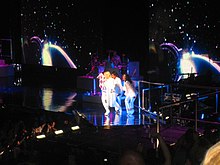 A blond woman in a white and purple leotard and a cape, which she opens up towards audience in front of her. She is flanked by two similarly dressed women.