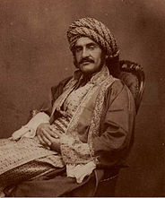 Sepia photograph of a man in 19th century Middle Eastern dress, with a large moustache, reclining in a chair with his hands crossed across his lap.