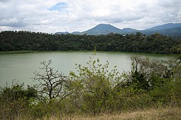 Circular volcano crater lake with sharp tree covered cliff banks and other volcanos in background.