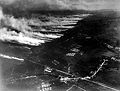 Vue d'avion en noir et blanc montrant des champs et un alignement de longues traînées de fumée blanche.