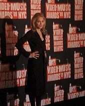 A blond woman standing in front of a black and red wall, wearing a black dress, and with her hands on her waist.