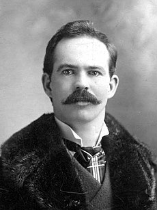 Upper body of a man with short hair and a thick moustache. He is wearing what appears to be a fur coat over a suit.