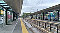 BelRed station on opening weekend in April 2024. The station has two platforms on the sides of the station with two tracks running down the center. There are shelters with glass walls and roofs along the platform, providing protection from the weather. The platform is made of concrete and has yellow lines and railings. There are benches for passengers to wait, and there are signs with information about the station and train schedules. The sky is cloudy, and there are puddles on the track bed.