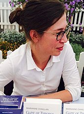 Photo d'une femme brune de profil (droit), appuyée des coudes sur une table, regardant sur la droite. Elle est coiffée d'un chignon, et porte des lunettes noires et une chemise blanche. Un jardinet clos est visible à l'arrière-plan, et des livres posés sur la table au premier plan, dont celui intitulé Harry Potter à l'école de la philosophie.