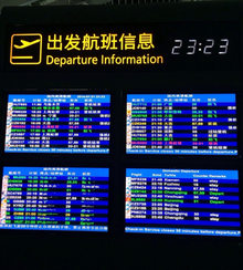 Four monitors with information on the departures of flights displayed in different colors of text on backgrounds of varying shades of blue. Three are in Chinese and one is in English. At the top is an icon with an airliner taking off and, in yellow, the words "Departure Information" in English and Chinese next to it. In the upper right is the time, 23:23, in white digital letters