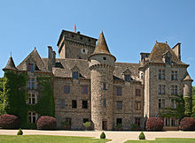 Sur cette photo, à l'arrière-plan sous un ciel d'un bleu pur, le donjon du château, d'une hauteur de 40 mètres, en forme de lourd parallélépipède quelque peu élargi tout en haut. Il date des débuts de la guerre de cent ans et domine le corps de logis, datant du XVIIe, mais transformé au XIXe selon la mode troubadour. L'aile gauche de celui-ci est une construction qui en fait partie, mais en ressort un peu avec son toit pyramidal avec, à l'extrême gauche, une tour ronde surmontée d'un toit pointu, moins élevée. À la droite du corps de logis, une construction plus détachée encore de l'ensemble tout en y demeurant liée aussi, plus haute, surmontée d'un toit pyramidal tronqué à quatre faces. Au centre une haute et plus grosse tour, plus détachée du reste, également au toit pointu, dépasse tout et même (mais par illusion d'optique), le donjon médiéval.