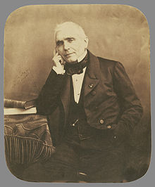 white man in late middle age, seated. He is clean shaven, and has white hair