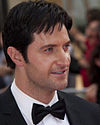 A dark-haired man wearing a jacket and bow-tie smiling at the audience off-camera.