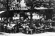 Vue en noir et blanc d'une terrasse de café bondée de consommateurs assis, au début du XXe siècle.