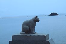 Photo couleur (couleur dominante = bleu) d'un sculpture en pierre d'un chat assis, vu de profil. Une étendue d'eau en arrière-plan.