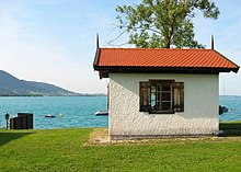 A small lakeside building showing a single window in a white wall below a sloping red roof