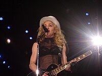 Picture of a middle-aged blond woman uptill the waiste, singing in front of a microphone. Her hair is in waves and falls upto her shoulders. She appears to be wearing a black bra covered with a sleeveless netted covering and wears a white hat on her head. There are black gloves on her hand and she plays an electric guitar. Behind her, to her left, a flood light is visible.