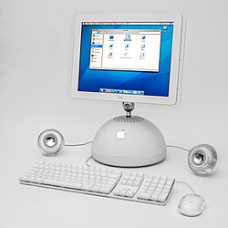 A white computer with a hemispherical base and shiny Apple logo, attached to a flat screen with a metal arm. A keyboard and mouse sit in front of the computer, while flanking the base are round, shiny speakers.