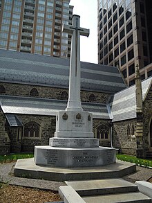 Photo en couleurs d'une grande croix posée sur un socle portant des inscriptions devant un bâtiment en pierres avec des gratte-ciels en arrière-plan