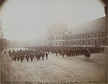 Photo en noir et blanc de militaires en uniforme en formation incluant une fanfare à la gauche de la formation principale et un militaire monté à cheval se tenant devant celle-ci