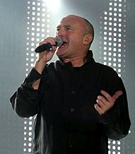 A man with a shaved head wearing black and singing into a microphone. The background contains dotted lighting on a stage.