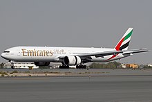 A mostly white Boeing 777, with some red, green and black markings, of Emirates, in flight, facing left.