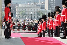 Photo en couleurs de militaires portant une tunique rouge et un bonnet à poil noir et tenant un fusil à la main se tenant debout en deux rangées de chaque côté d'un tapis rouge ; derrière, des militaires porant une tunique verte foncée et un kilt de tenant debout en deux rangées derrière un corbillard dont la porte de derrière est ouverte ; en arrière-plan, des bâtiments d'un centre-ville