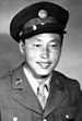 Head and shoulders of a smiling young man wearing a peaked cap and, over a shirt and tie, a military jacket with a round pin on each lapel.