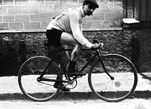 Photographie en noir et blanc d'un cycliste sur sa bicyclette, les mains tenant le guidon, la jambe gauche à terre