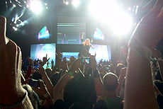 A long distance image of a stage, with five video screens, and floodlights falling from above. In the middle, a blond woman stands, with her right hand up in the air. At the front, the back-profile of numerous audience members, with their hands outstretched, can be seen.