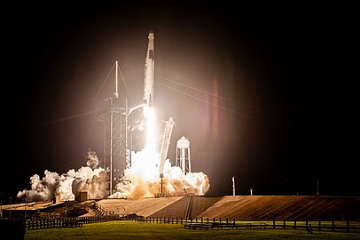 A tall white rocket lifts off from a launch pad, the rockets base glistening in its flame, as a plume of smoke forms at the launch pad