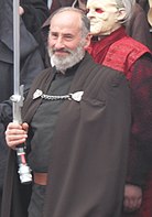 Homme âgé, avec des cheveux gris, portant une toge sombre et un sabre laser incurvé, souriant.