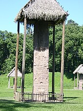 A tall, narrow monument covered by a thatched roof. Two further stelae are visible in the background, also covered by thatched roofs, against a backdrop of trees.