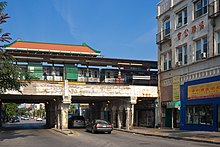 Road with a railroad bridge crossing it. A train waits at a station on the bridge as passengers disembark