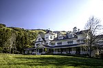 A white mansion house topped by a black roof stands amid green hills and forests.