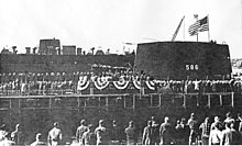 A large number of people are gathered around a platform erected just in front of a submarine.