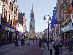 Falkirk High Street with the Falkirk Steeple dominating the centre of the picture. Shops to the left and right are visible with many shoppers on the pedestrianised street.