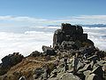 Photo couleur d'un amoncellement de rochers gris au sommet d'une montagne. L'arrière-plan est composé d'une nappe de nuages blancs, sous un ciel bleu nuageux.