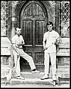 Two schoolboys holding racquets racquets, standing on wooden steps either side of an arched wooden double door to a school building