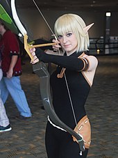 Cosplay d'une femme, en habit moulant noir, cheveux blonds et oreilles pointues. Elle bande un grand arc, prête à tirer.