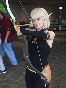 Cosplay d'Evangelyne, en habit moulant noir, cheveux blonds et oreilles pointues. Elle bande un grand arc, prête à tirer.