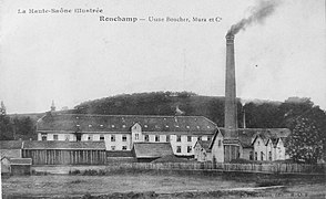 Photo noir et blanc montrant un long bâtiment industriel anciens surmonté d'une cheminée fumante et une petite colline proche.