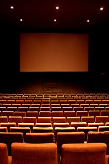 Salle de cinéma, avec plusieurs rangées de fauteuils rouge et un écran géant blanc en fond.