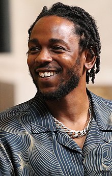 Lamar in a dress shirt and prominent metal necklace looks to his right and smiles.