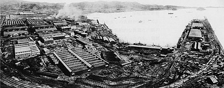 Photo aérienne d'un complexe industriel en bord de mer. De très nombreux hangars sont visibles, desquels par endroits s'échappe de la vapeur. Le long de la mer, est aménagé un quai pour charger et décharger des marchandises.