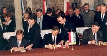 Three men in dark suits sit at a table covered in a red tablecloth signing documents, around them stand others in dark suits.